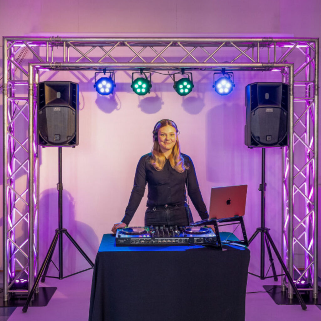 DJ seisoo valaistun trussikaaren alla DJ-laitteiden ja kannettavan tietokoneen takana. Molemmin puolin on suuret kaiuttimet, yläpuolella neljä värikästä spottivaloa. Taustalla violetti tunnelmavalaistus.